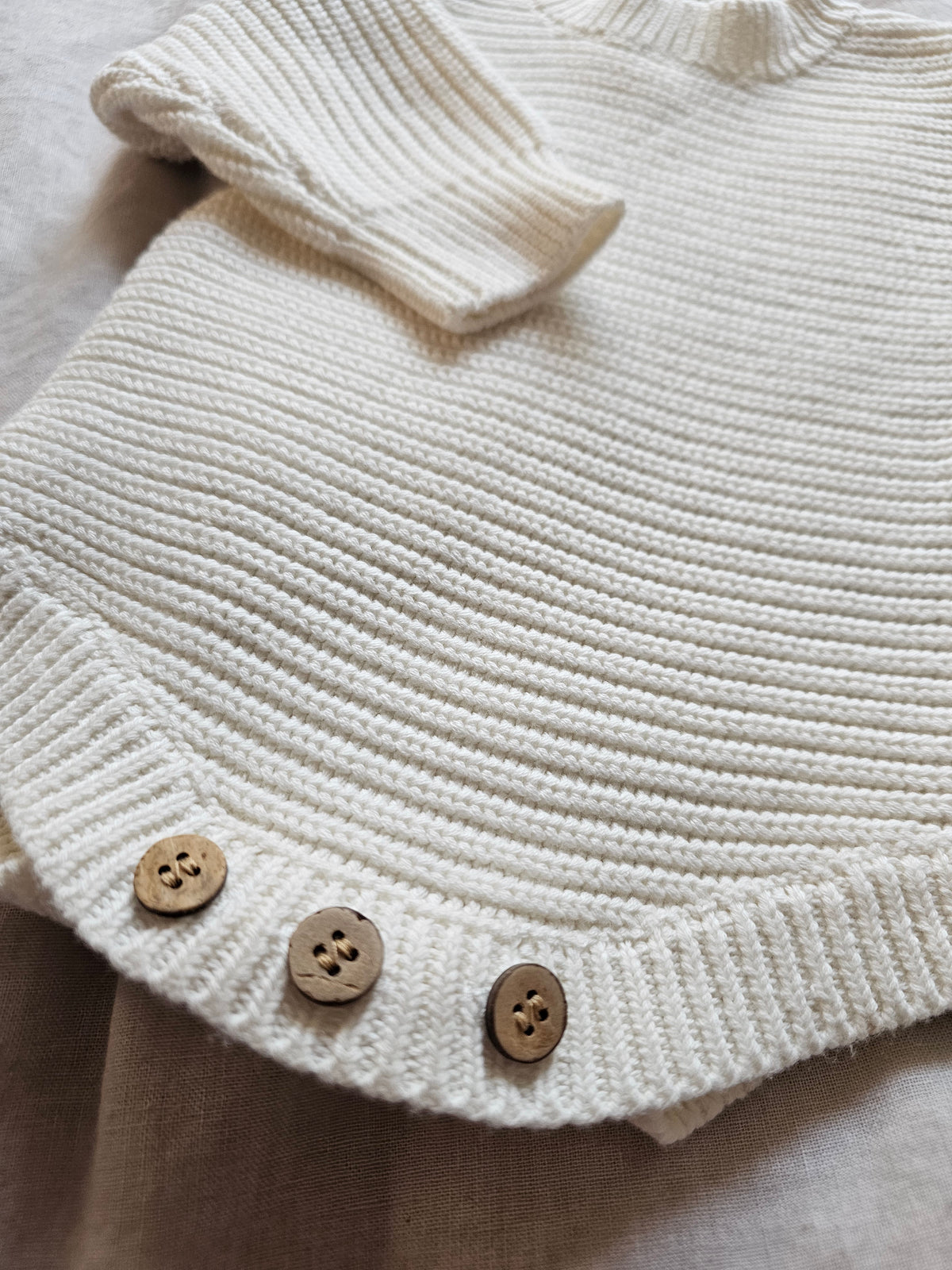 Close-up of a cream-colored ribbed knit garment,with three wooden buttons along the hem, laid flat on a neutral fabric background.