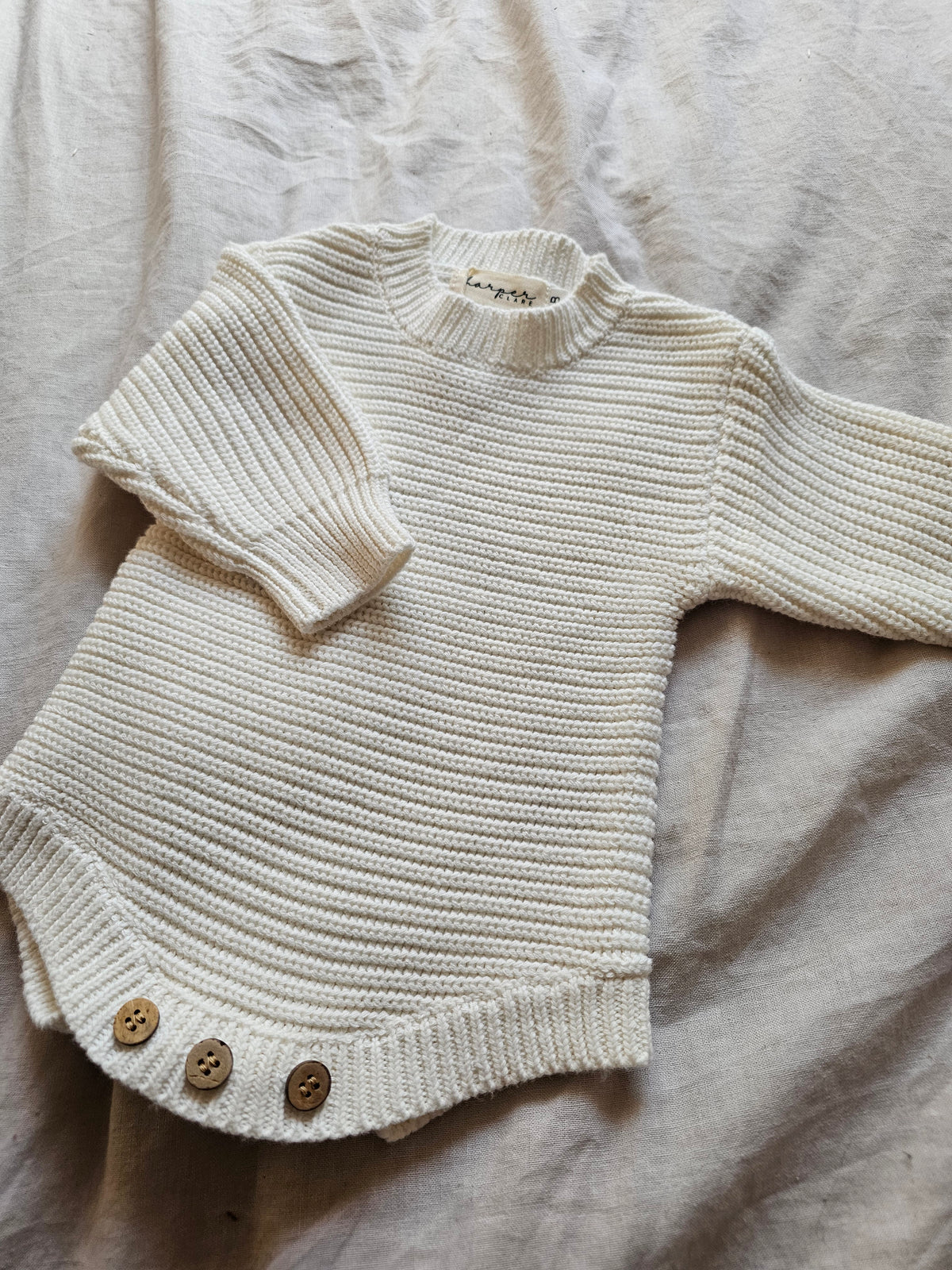 white ribbed knit baby romper with long sleeves and three brown buttons at the crotch, laid flat on a textured beige fabric background.