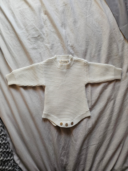white ribbed knit baby romper with long sleeves and three metal snap buttons at the crotch, laid flat on a beige fabric background.