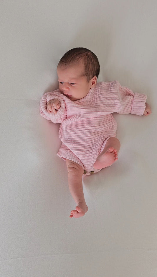 A newborn baby lying on their side wearing a pink ribbed long-sleeve bodysuit, photographed from a top-down angle on a plain light background.