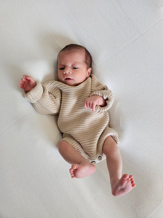 A newborn baby lying on their back on a white blanket, wearing a beige chunky-knit long-sleeve romper.