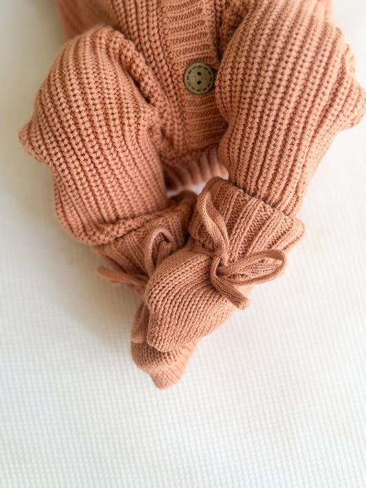 A close-up of a terracotta knitted baby romper with wooden buttons paired with matching knit booties, laid flat on a white textured fabric background.