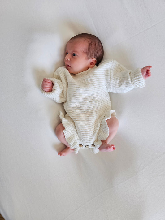 A newborn baby lying on a white blanket, wearing a cream-colored ribbed knit romper with ruffled legs and three front buttons.