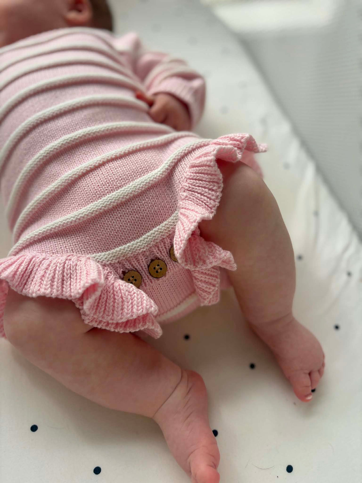 A close-up lifestyle shot of a baby wearing a pink and white striped knit bodysuit with ruffled trim and decorative buttons.