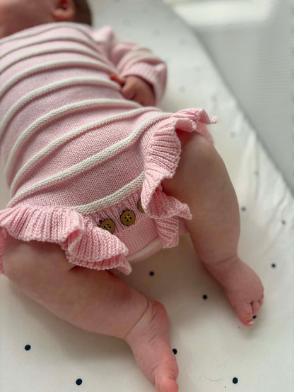 A close-up lifestyle shot of a baby wearing a pink and white striped knit bodysuit with ruffled trim and decorative buttons.